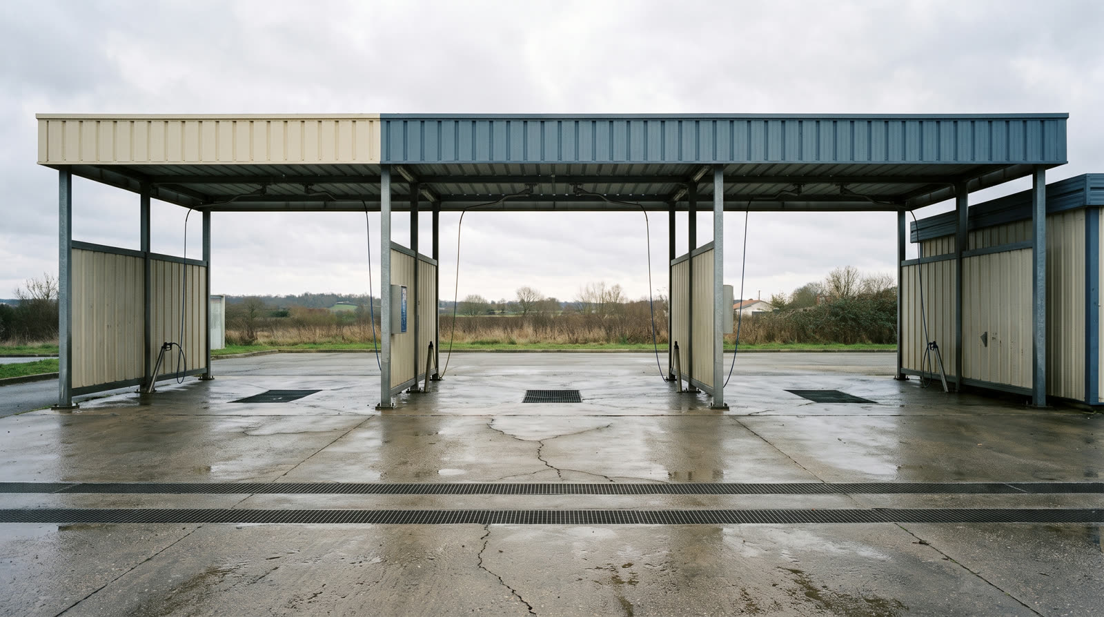 Station de lavage automobile en libre-service : pistes couvertes sous auvent métallique, dalle béton et caniveaux — surfaces secondaires couvertes à pondérer distinctement.